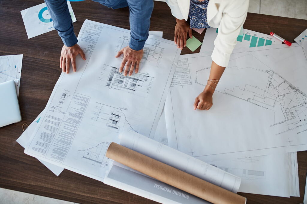 Overhead view of two people reviewing architectural blueprints and floor plans spread across a table, with sticky notes and documents, illustrating the home design selection process