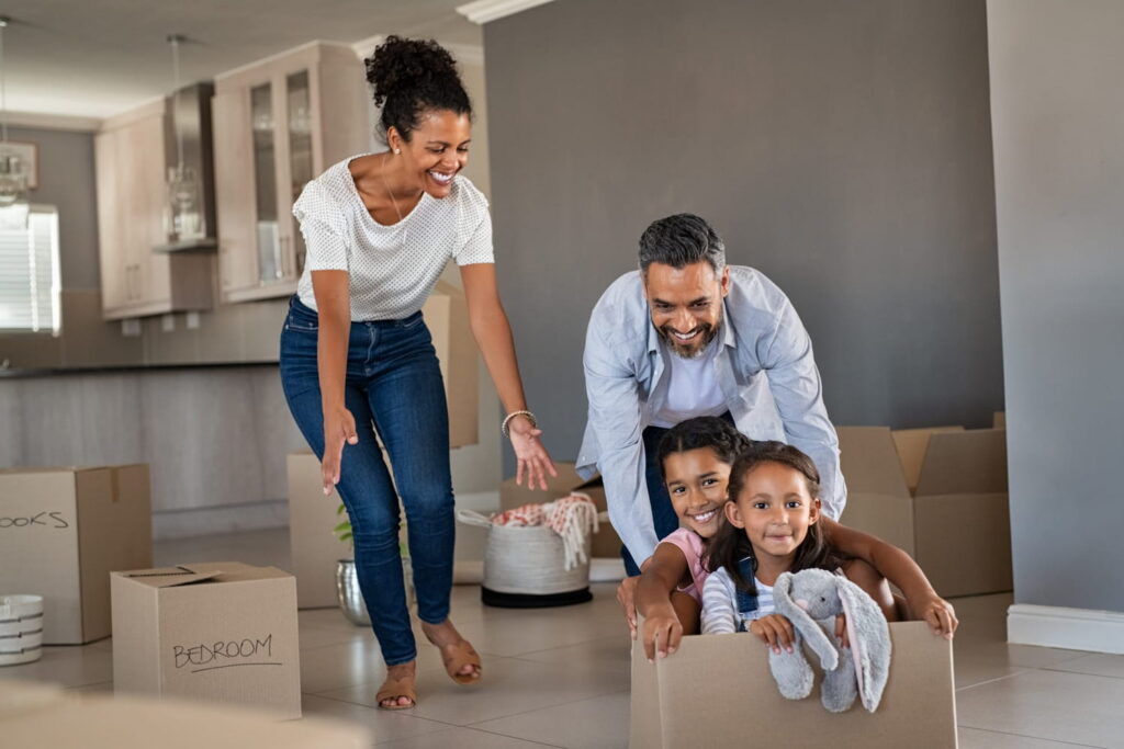 A smiling family of four celebrating move-in day in their new Edmonton Pacesetter home; a father pushes two young daughters in a moving box while the mother cheers them on in a modern open-concept kitchen and living area.